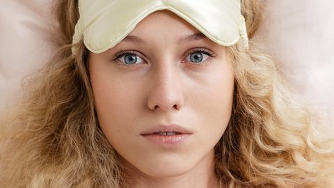Blonde woman wearing a sleep mask, looking awake and worried, highlighting insomnia.