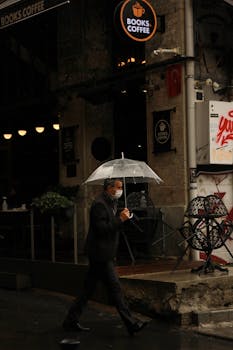 An adult male with an umbrella passing by a cozy urban cafe during a rainy day, with a masked face.