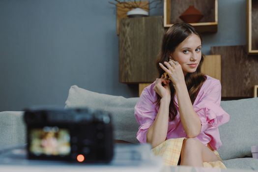 Caucasian woman in pink blouse adjusting earrings, sitting on a sofa with camera in foreground.