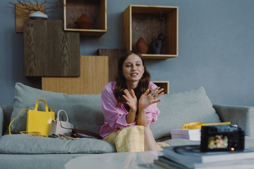 Free Young woman sitting on a couch, recording a fashion video blog indoors with stylish accessories and camera. Stock Photo