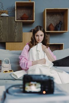 Woman with pink sleeves and white coat sits on couch during filming setup, serious expression.