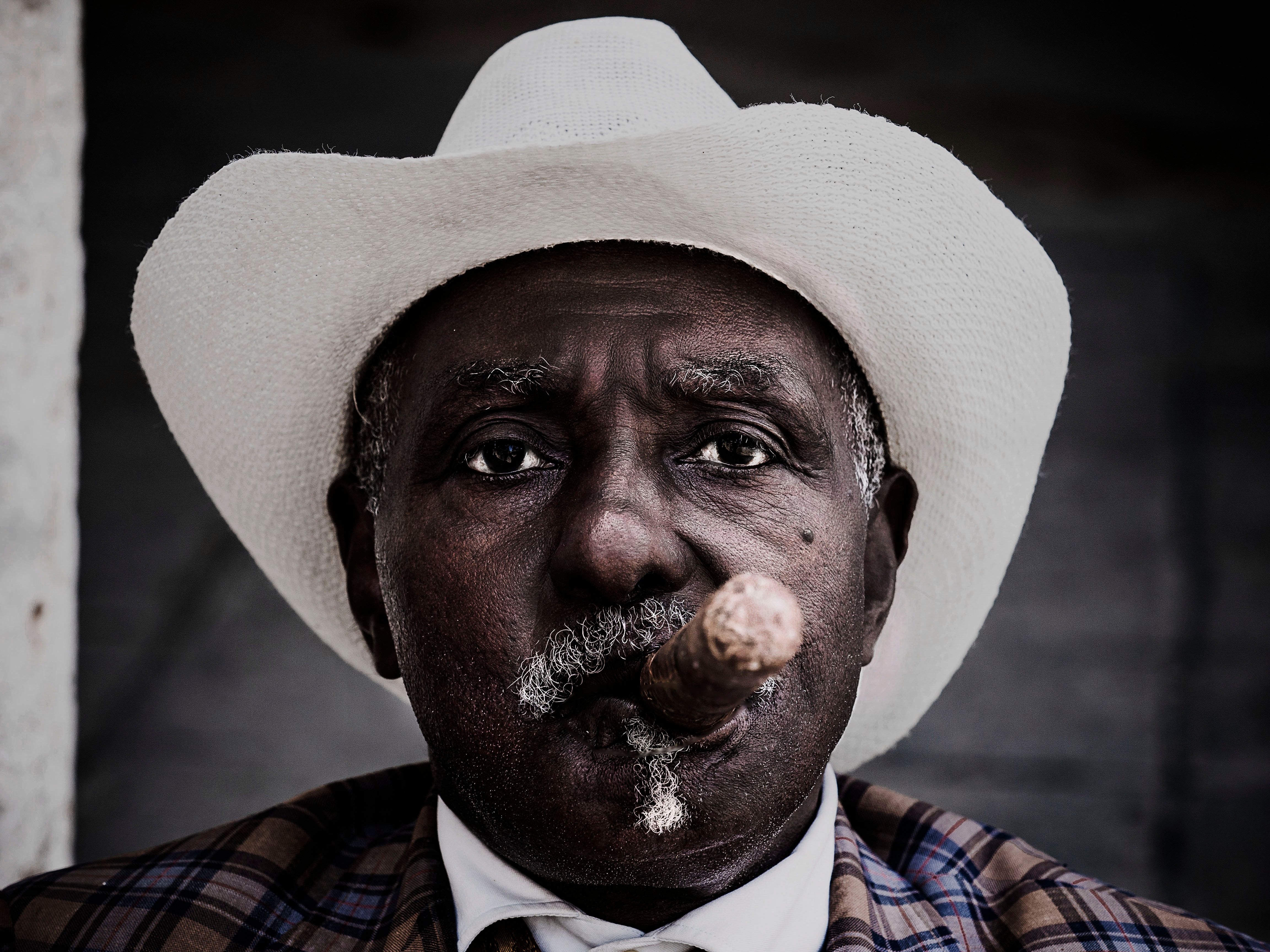 An expressive close-up portrait of a man smoking a cigar, wearing a white cowboy hat.