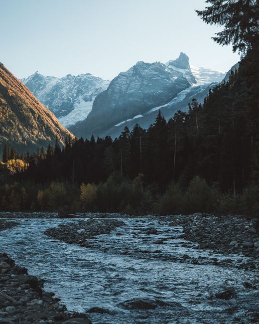 Breathtaking view of Dombay's snow-covered mountains with a flowing river and dense forest.