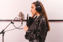 Young woman in leather jacket singing into microphone in studio setting.