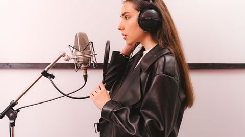 Young woman in leather jacket singing into microphone in studio setting.