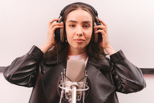 Woman in a leather jacket and headphones singing into a microphone in a studio.