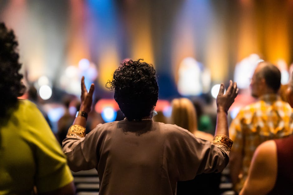 A woman with curly hair raises her hands in a colorful, vibrant concert setting filled with energy.