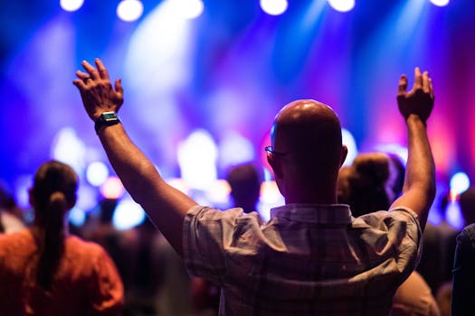 Bald man from behind raising hands in celebration at a vibrant live concert, colorful lighting.