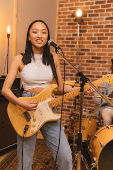 Young Asian woman singing and playing electric guitar in a music studio with a drum set in the background.