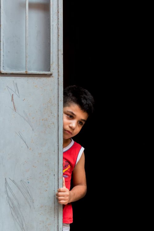 Free A young boy in a red sleeveless shirt peeks out from a metal door against a dark background. Stock Photo