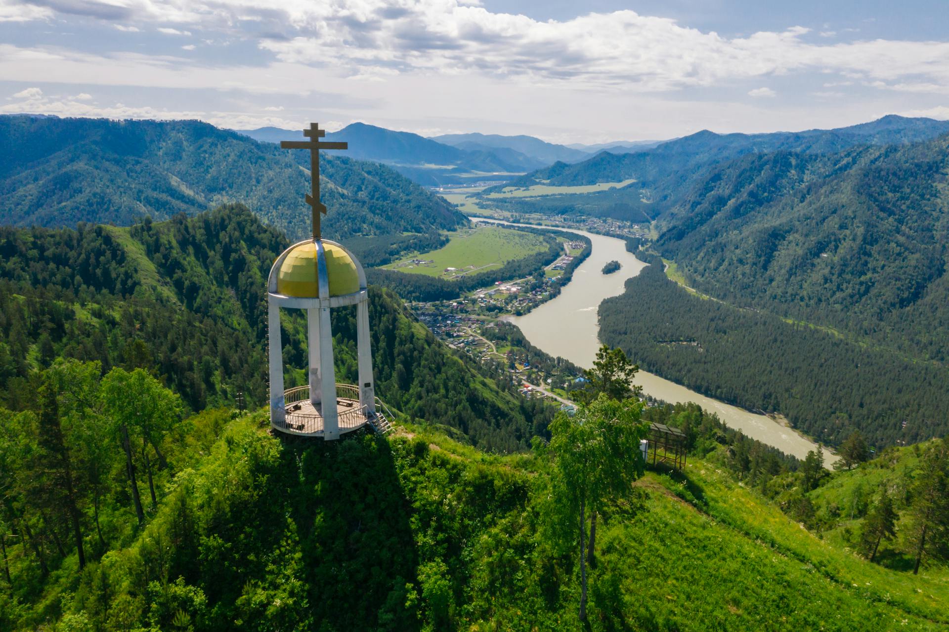 Stunning aerial view of a river valley with a cross monument and lush green mountains.