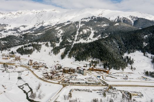 Drone shot of a snowy mountain ski resort with coniferous trees and snowy peaks.