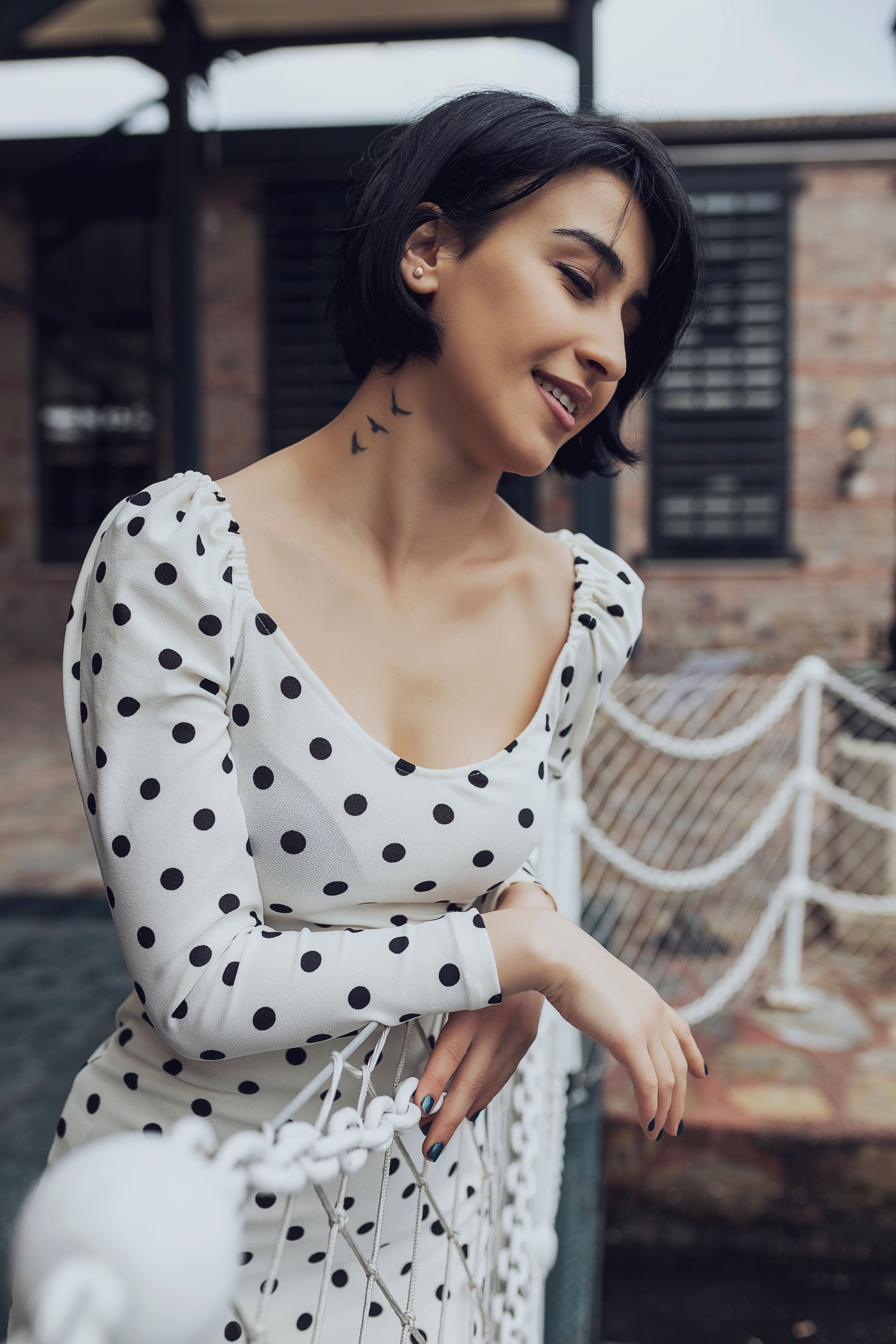 Elegant woman with short hair in polka dot dress smiling outdoors by a fence.