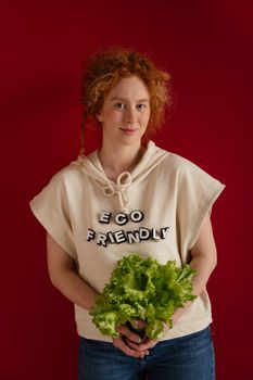 Red-headed woman holding lettuce against a red background wearing an 'eco-friendly' shirt.