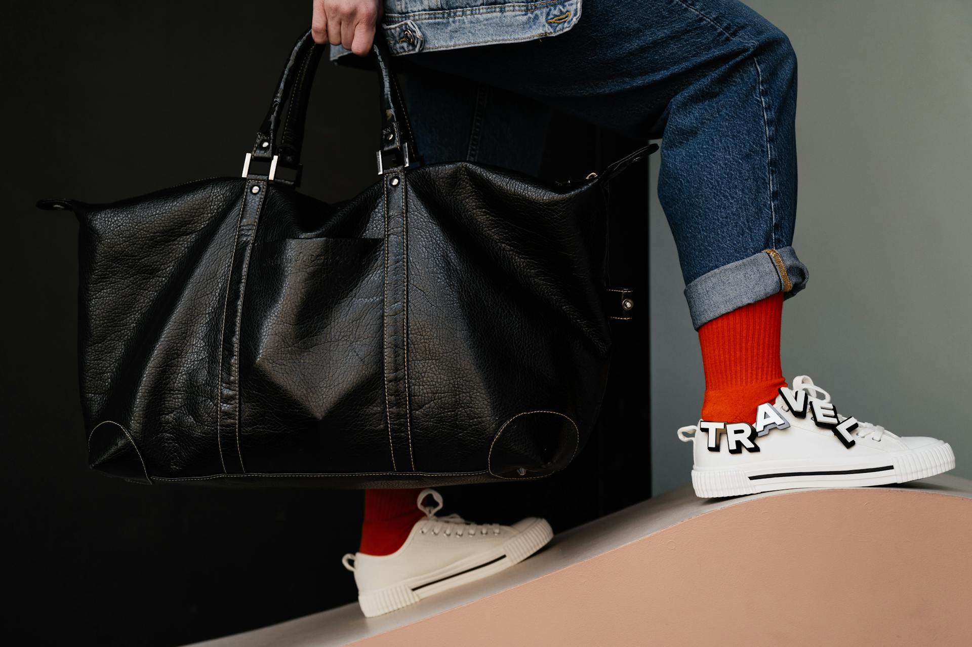 Close-up of person holding leather duffel bag with travel-themed sneakers. Trendy outdoor travel concept.