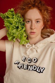 Portrait of a redhead woman in an eco-friendly hoodie holding lettuce against a red background.