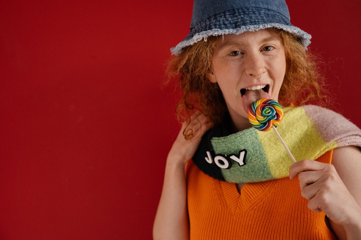 Woman in denim hat licking a colorful lollipop with joy on scarf.