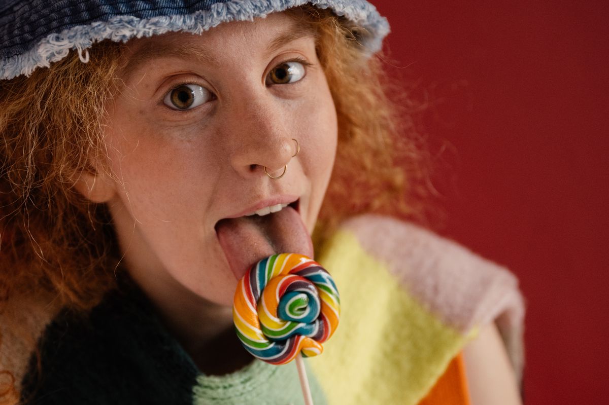 A playful woman with red hair enjoying a vibrant lollipop, expressing joy and silliness.