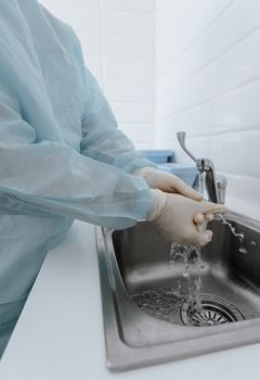 A healthcare worker in protective gear washing hands in a clinical setting sink.
