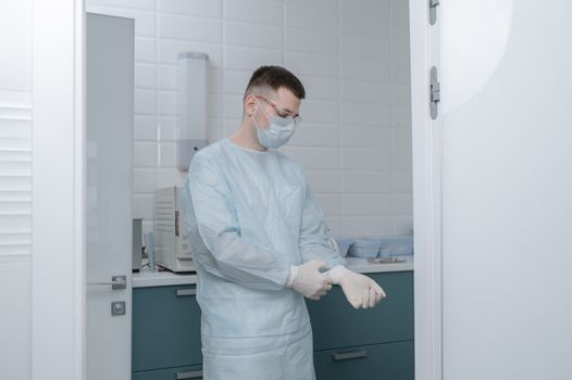 A healthcare professional in a lab coat and mask puts on gloves in a hospital room.