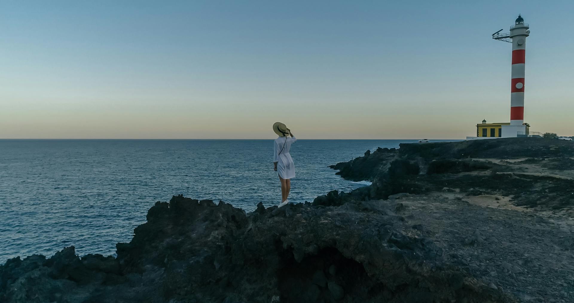 Woman in a white dress stands on a cliff by the lighthouse with ocean views at sunset.