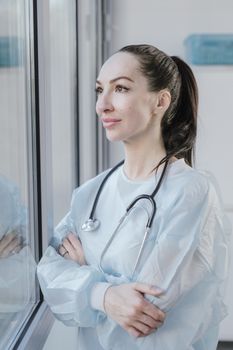 A female medical professional in a disposable gown looks thoughtfully out a window, stethoscope around her neck.