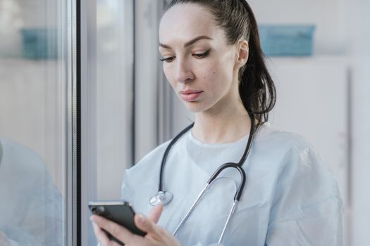 Portrait of a female doctor in a disposable gown using a smartphone in a hospital environment.