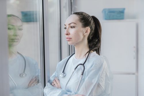 Free Female doctor in a lab coat with stethoscope, pondering while looking out a window. Stock Photo