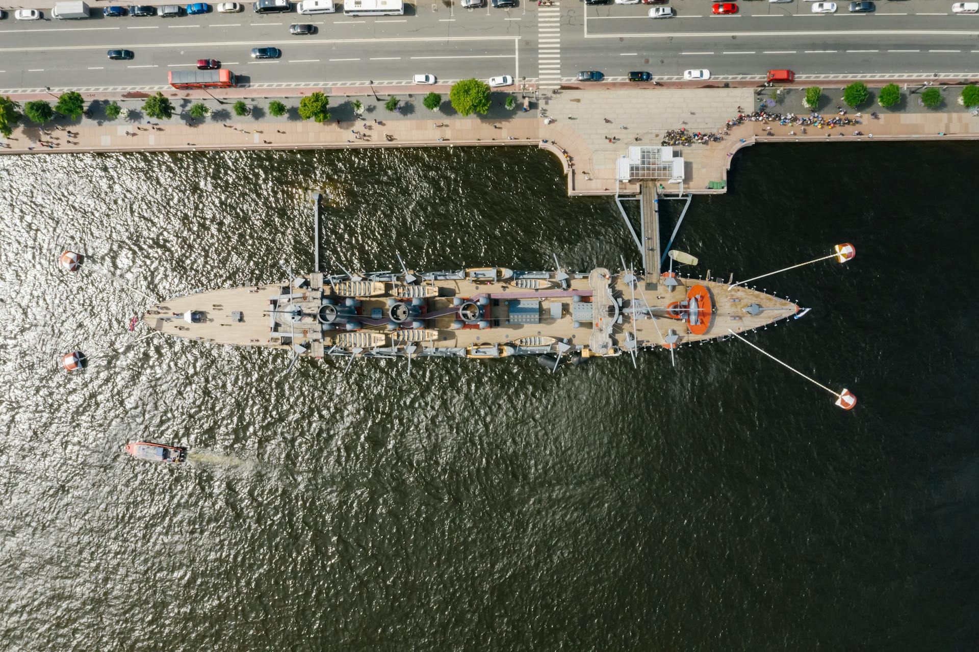 Top-down drone shot of a docked battleship with waterfront and adjacent road.