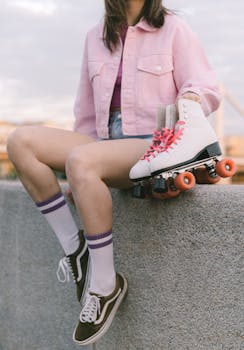 Young woman in a fashionable outfit holding roller skates while sitting outdoors on a concrete ledge.