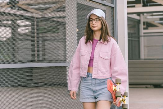 Young woman with skateboard wearing pink jacket and denim shorts walking outdoors. Trendy urban style.