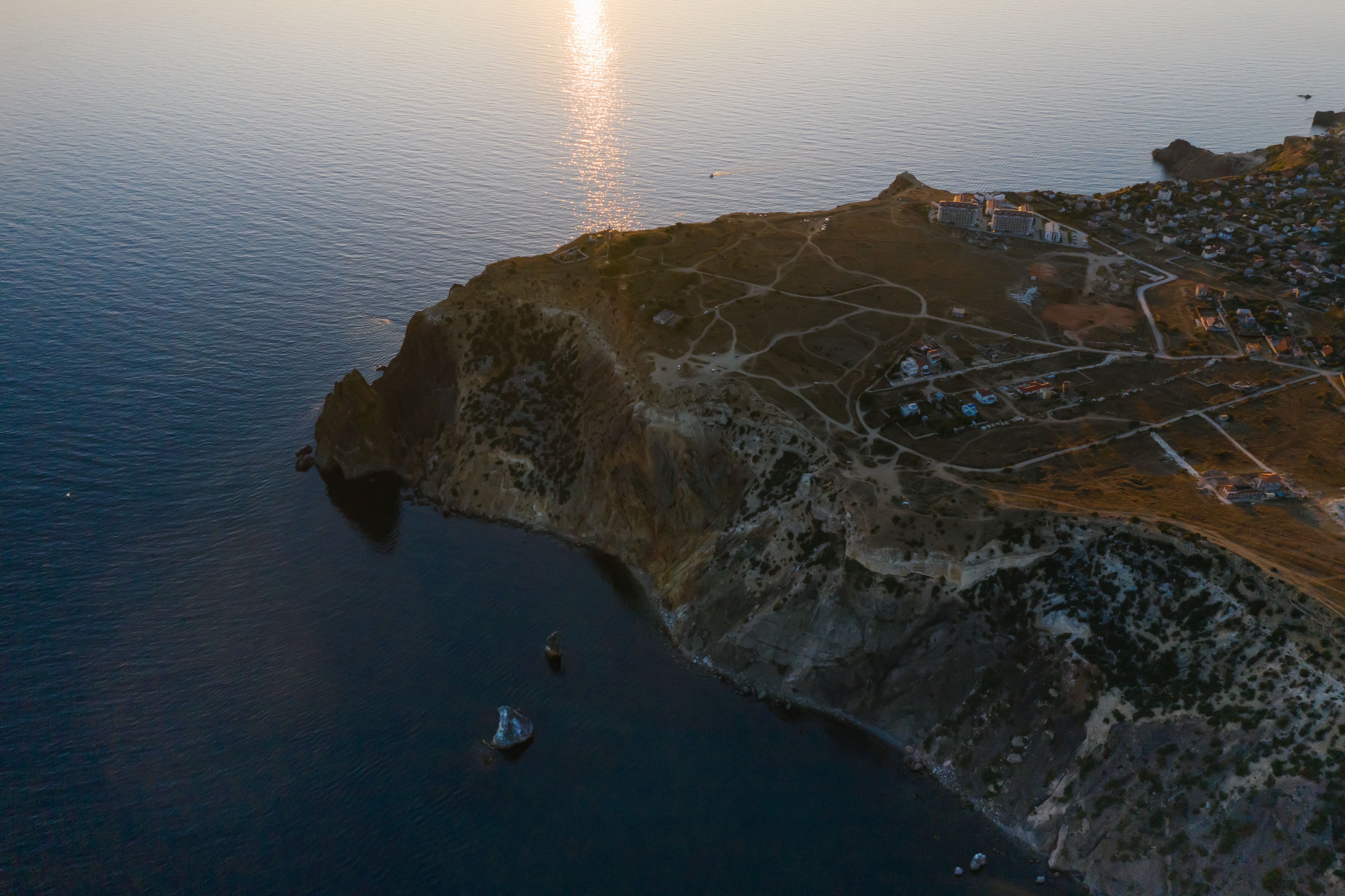 Aerial view of rugged coastal cliffs with winding paths and scattered buildings overlooking the sea at sunset