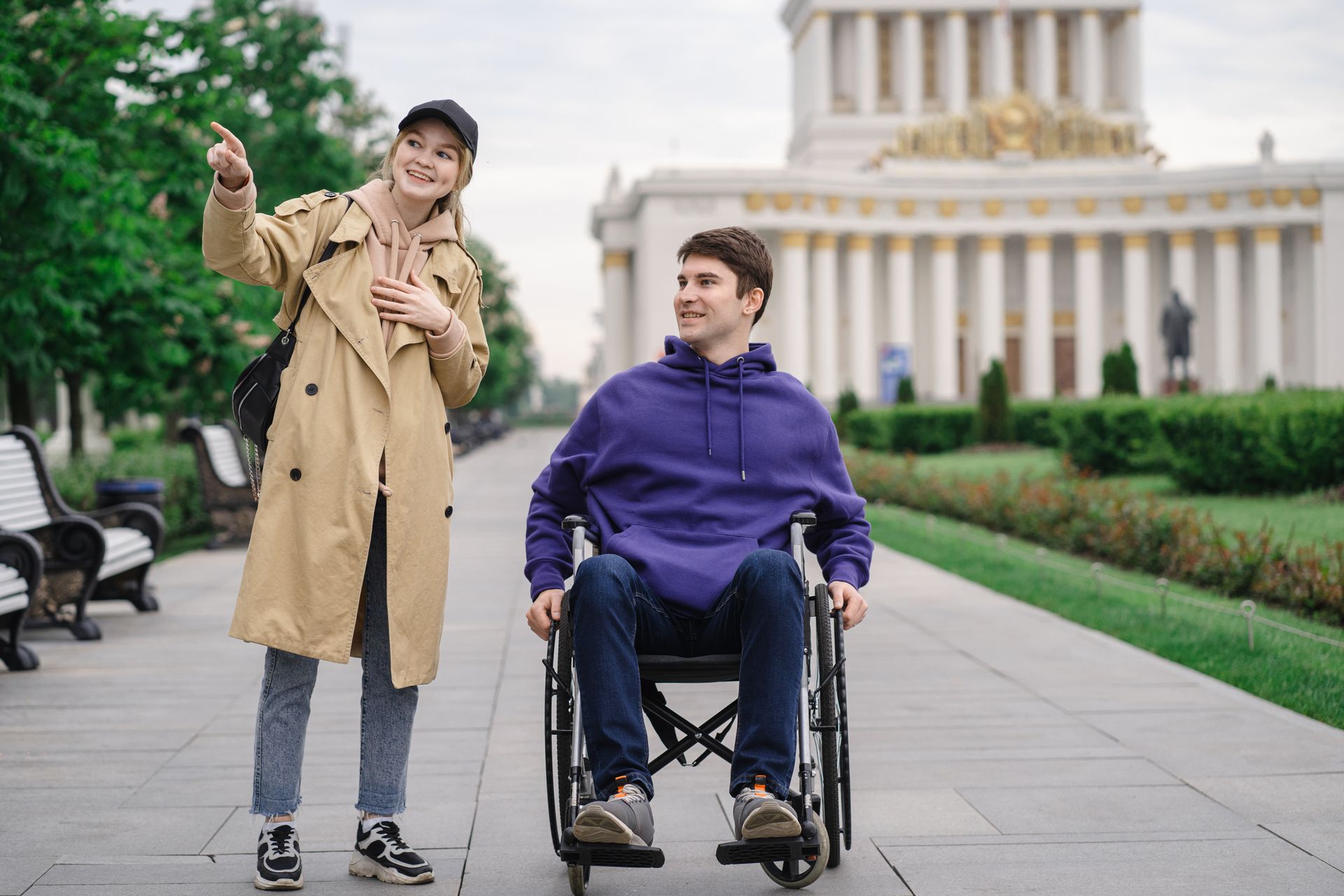 Woman pointing while man in wheelchair smiles outdoors by a historic building.