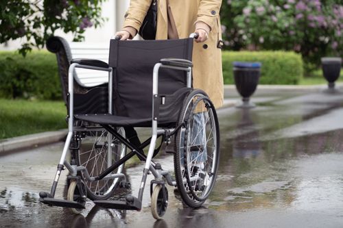 Free A person in a trench coat pushing an empty wheelchair along a wet outdoor path. Stock Photo