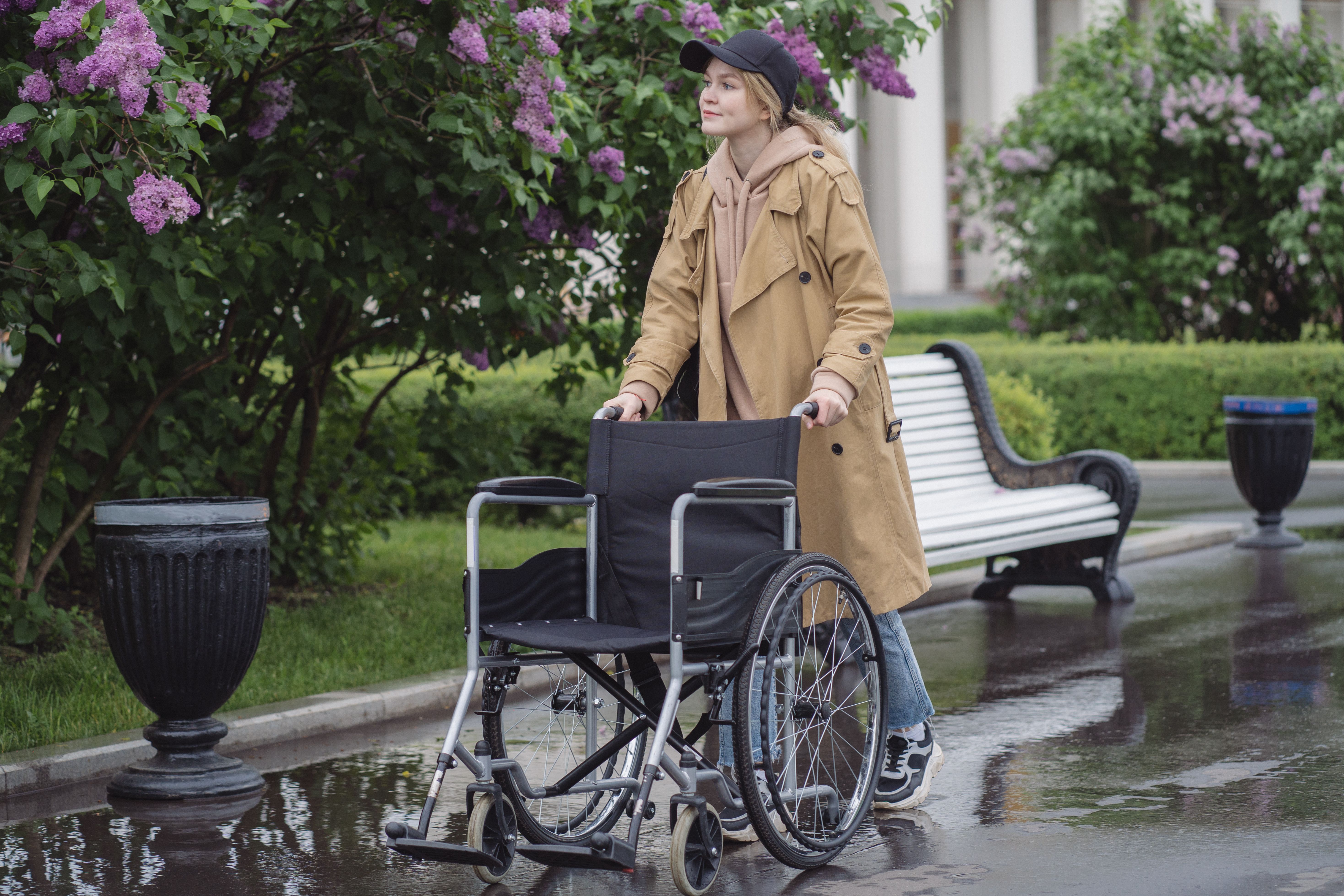 Woman in a trench coat and cap walking with an empty wheelchair on a wet path.