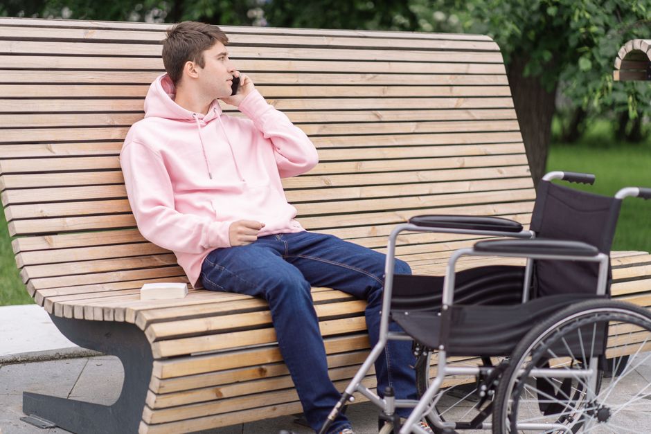 Casual young man in pink hoodie sitting on a bench talking on phone with wheelchair nearby.