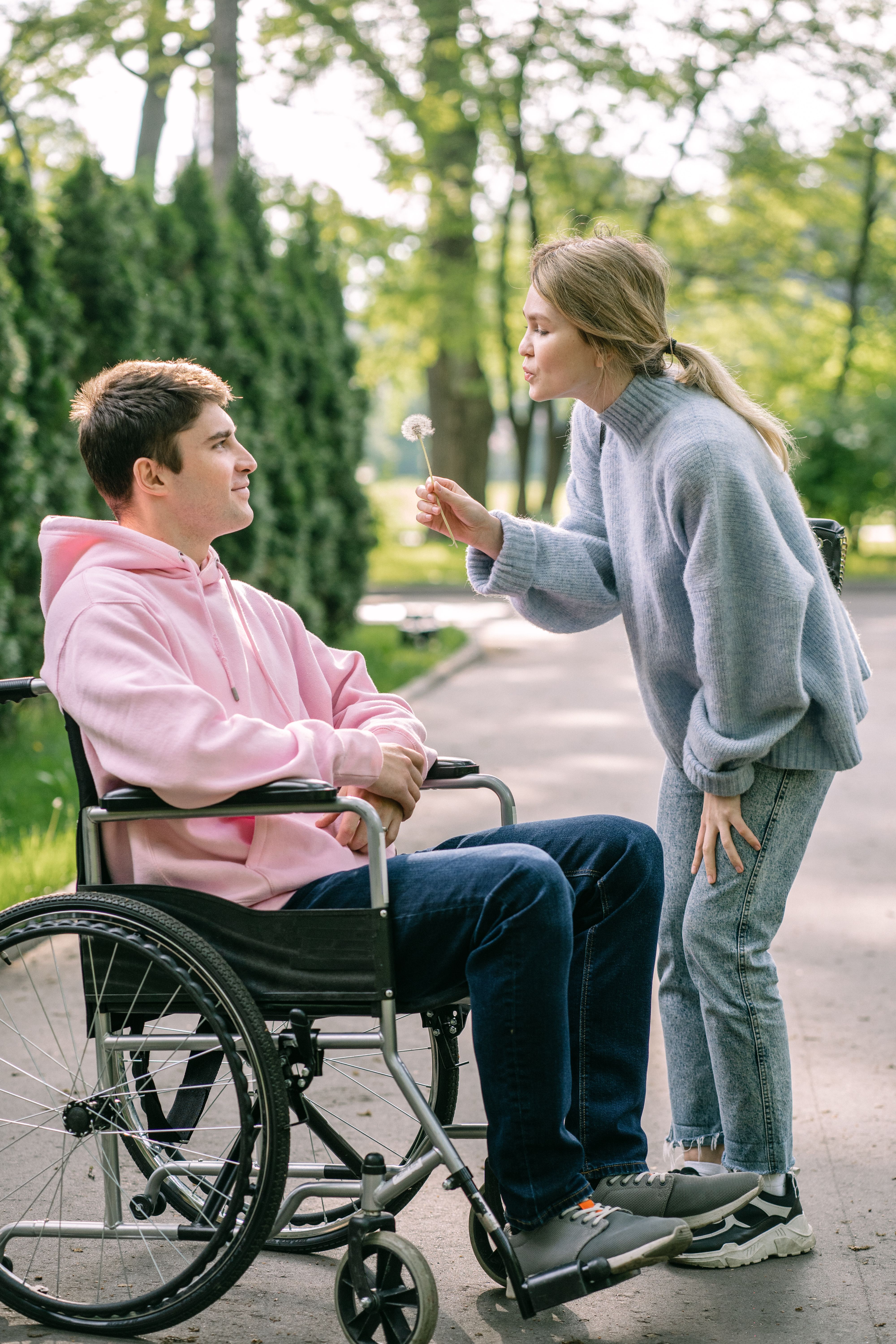 A young couple in a park, one in a wheelchair, laughing as dandelion seeds float around them