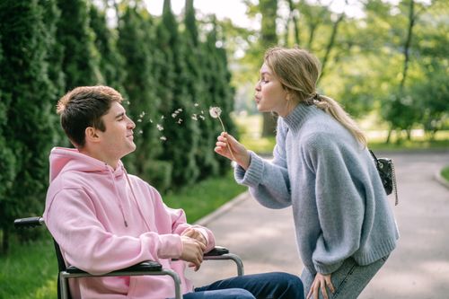 Free A woman blows a dandelion while a man in a wheelchair smiles, enjoying a sunny day outdoors. Stock Photo
