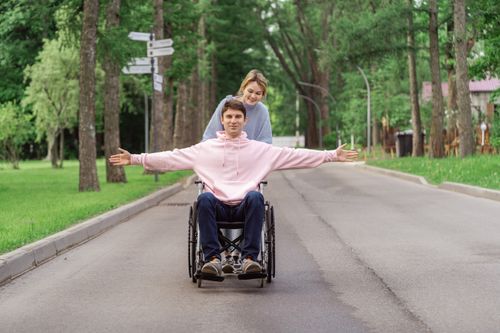 Free A young couple joyfully spends time outdoors, the man in a wheelchair being pushed by a woman. Stock Photo