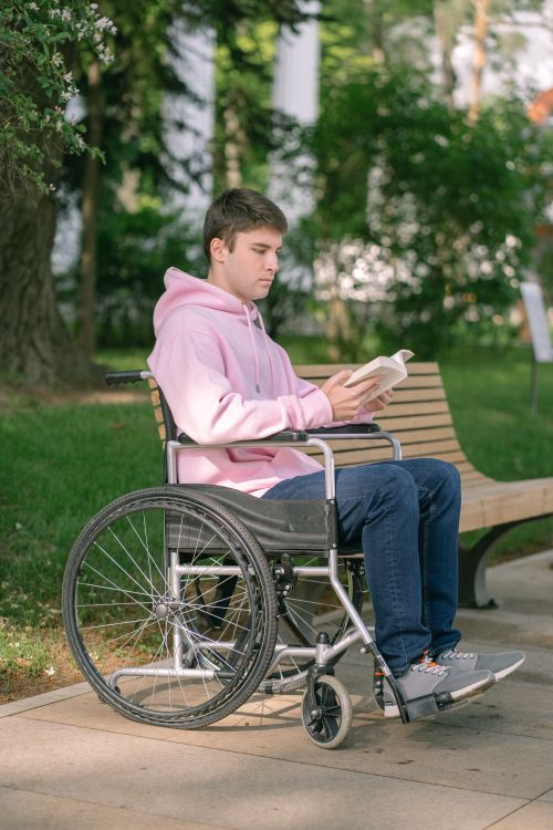 Free A young man in a wheelchair reading a book outdoors, wearing a pink hoodie on a sunny day. Stock Photo