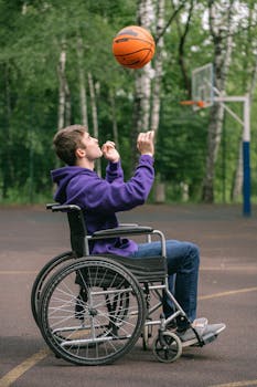 A young man in a purple hoodie plays basketball from his wheelchair on an outdoor court.