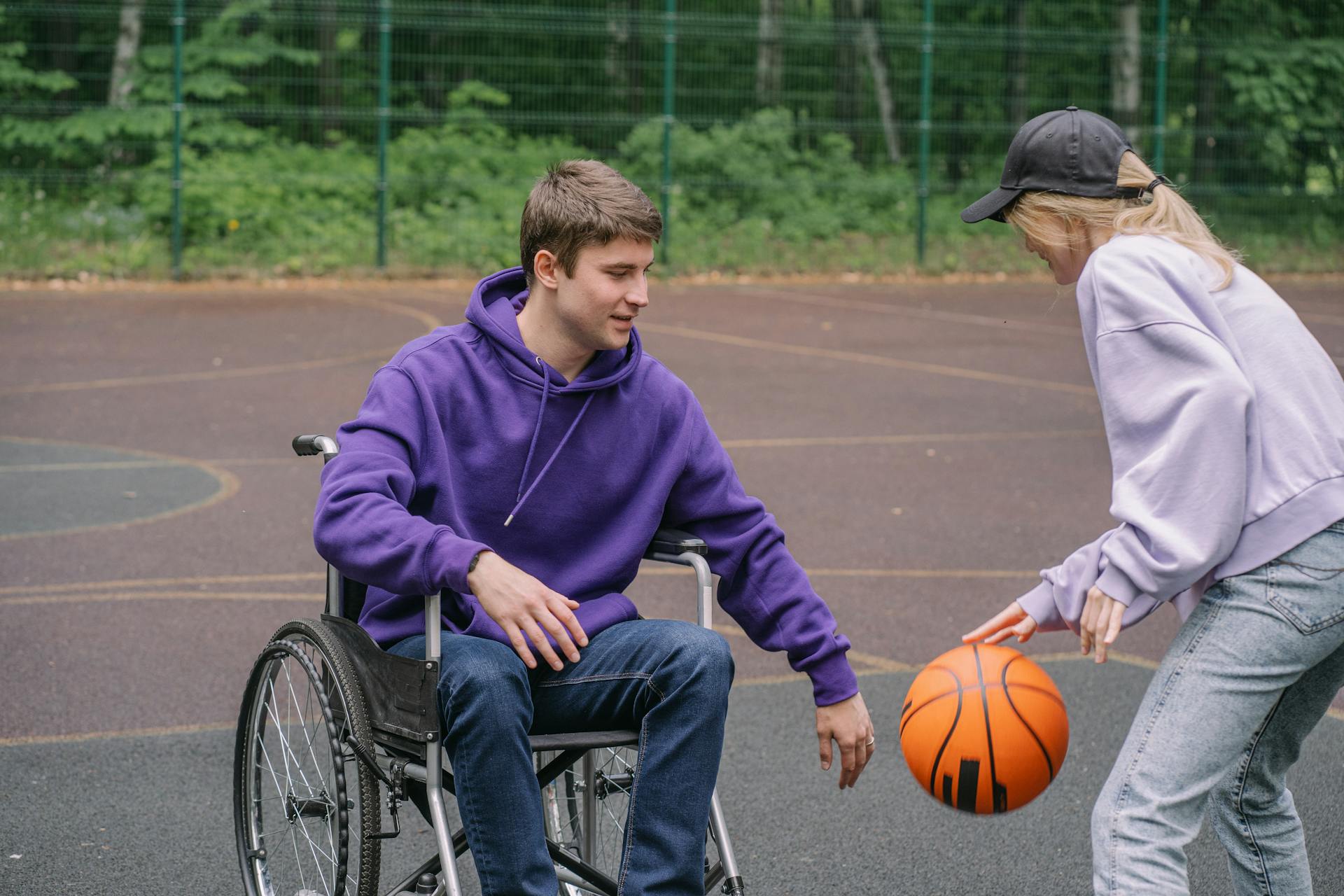 A man in a wheelchair plays basketball outdoors with a friend, promoting inclusion and sports.