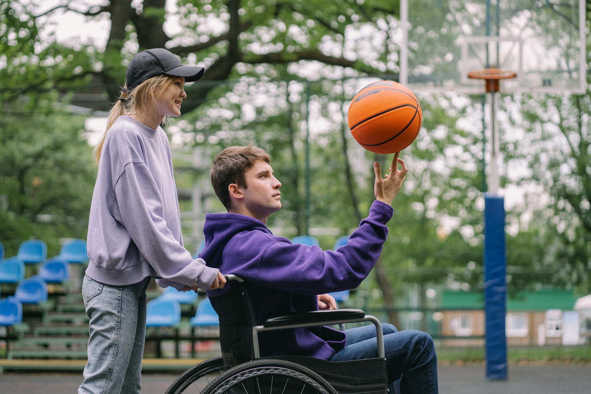 A young man in a wheelchair spins a basketball outdoors with a supportive woman by his side.