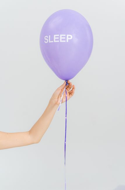 A minimalist photo featuring a hand holding a purple balloon with the text 'SLEEP' against a white background.