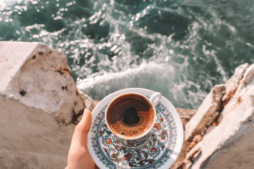 Free Hand holding ornate coffee cup by ocean waves. Serene outdoor scene with intricate design. Stock Photo