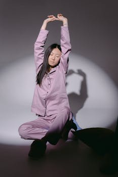 A woman in purple sleepwear stretches with eyes closed, sitting on the floor under soft lighting.