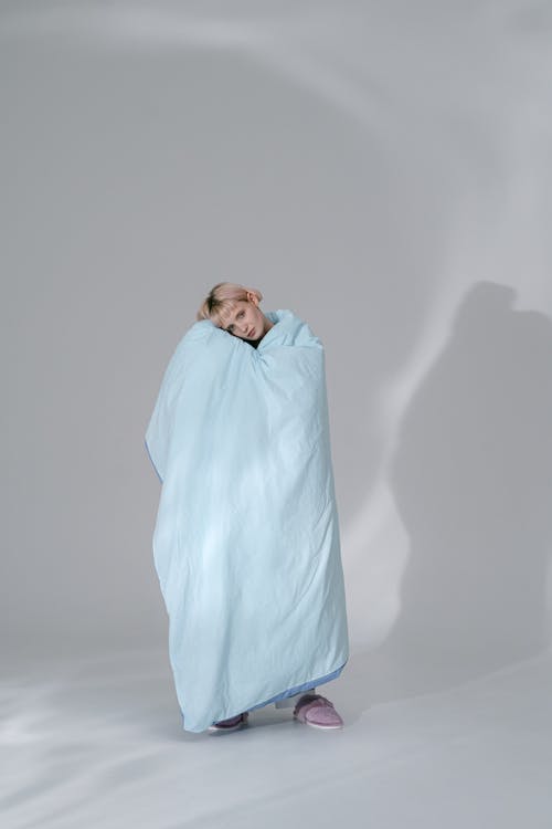 A Woman Wrapped with Blanket Standing Near White Wall while Looking at the Camera
