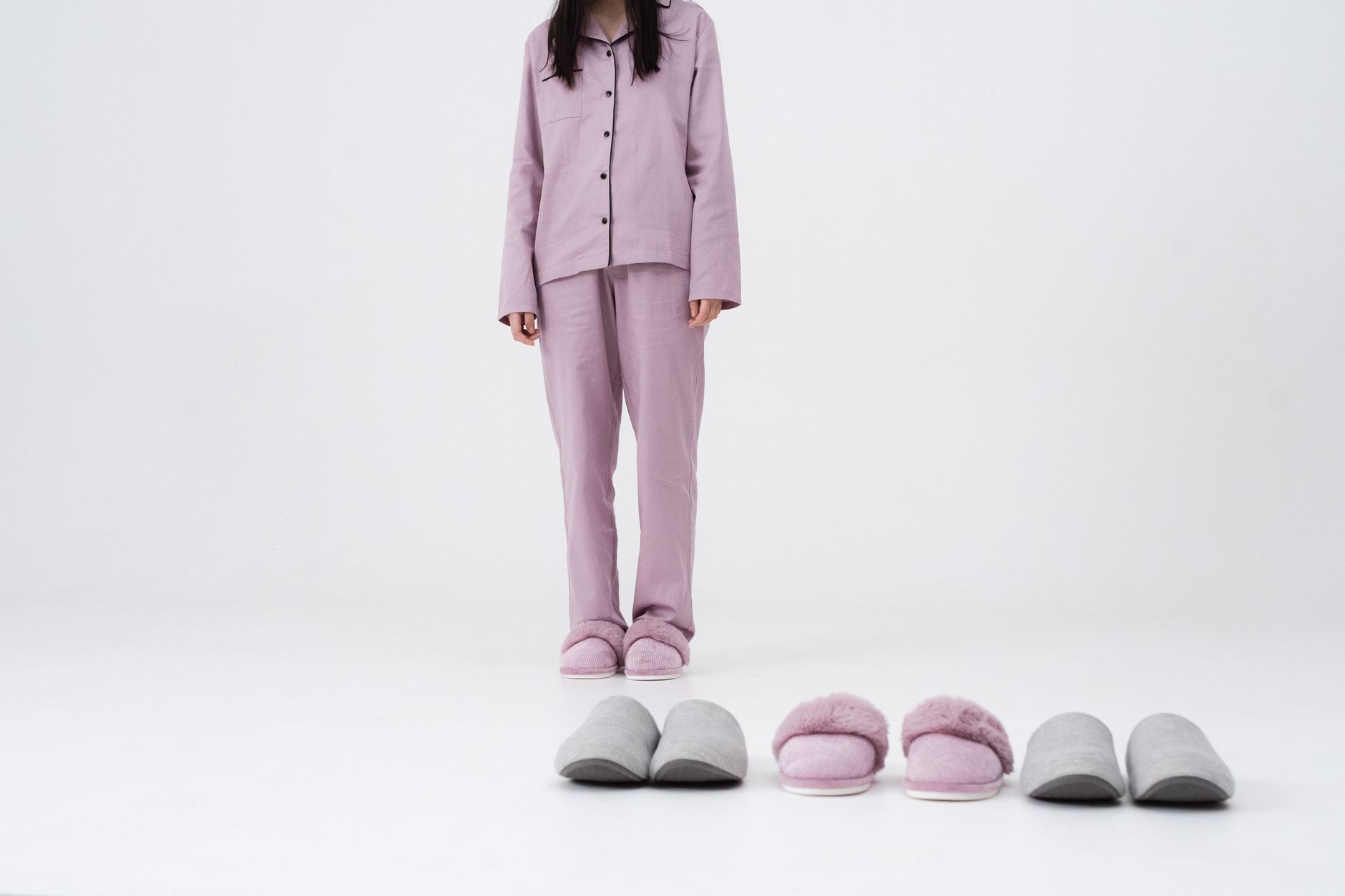 Stylish woman in pink pajamas standing with multiple slippers in a studio setting.