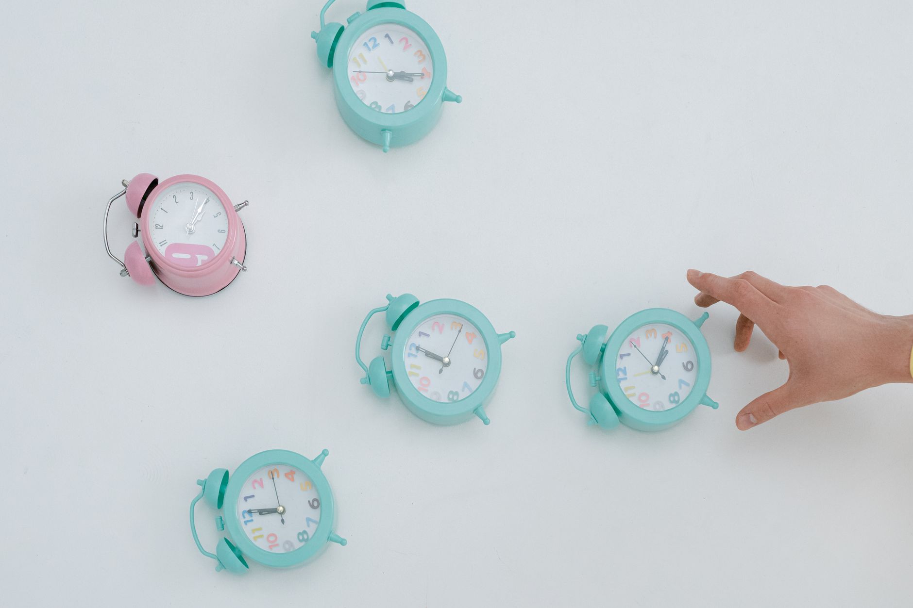 A hand reaching for pastel alarm clocks on a plain white background. Minimalist style.