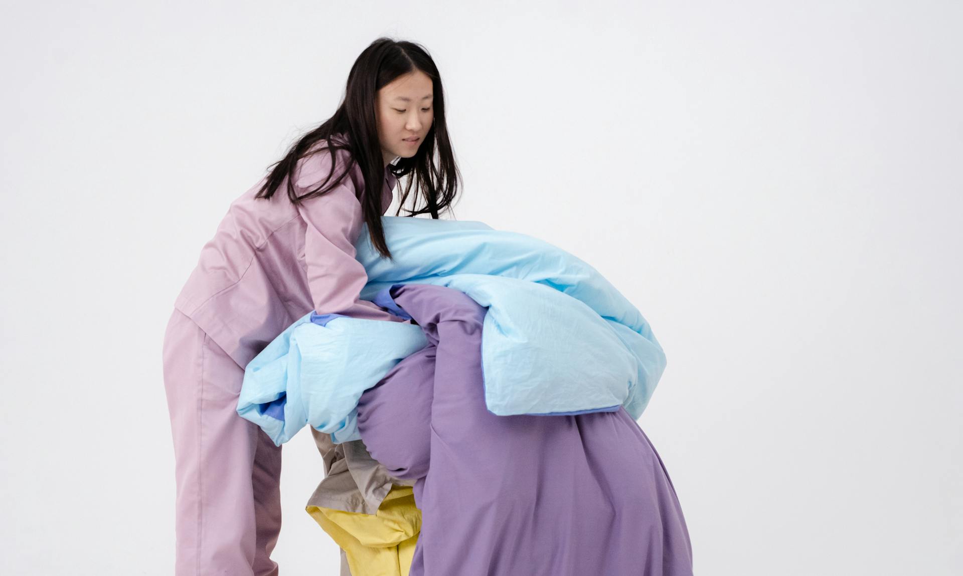 A young woman in purple pajamas arranging colorful blankets against a white background.